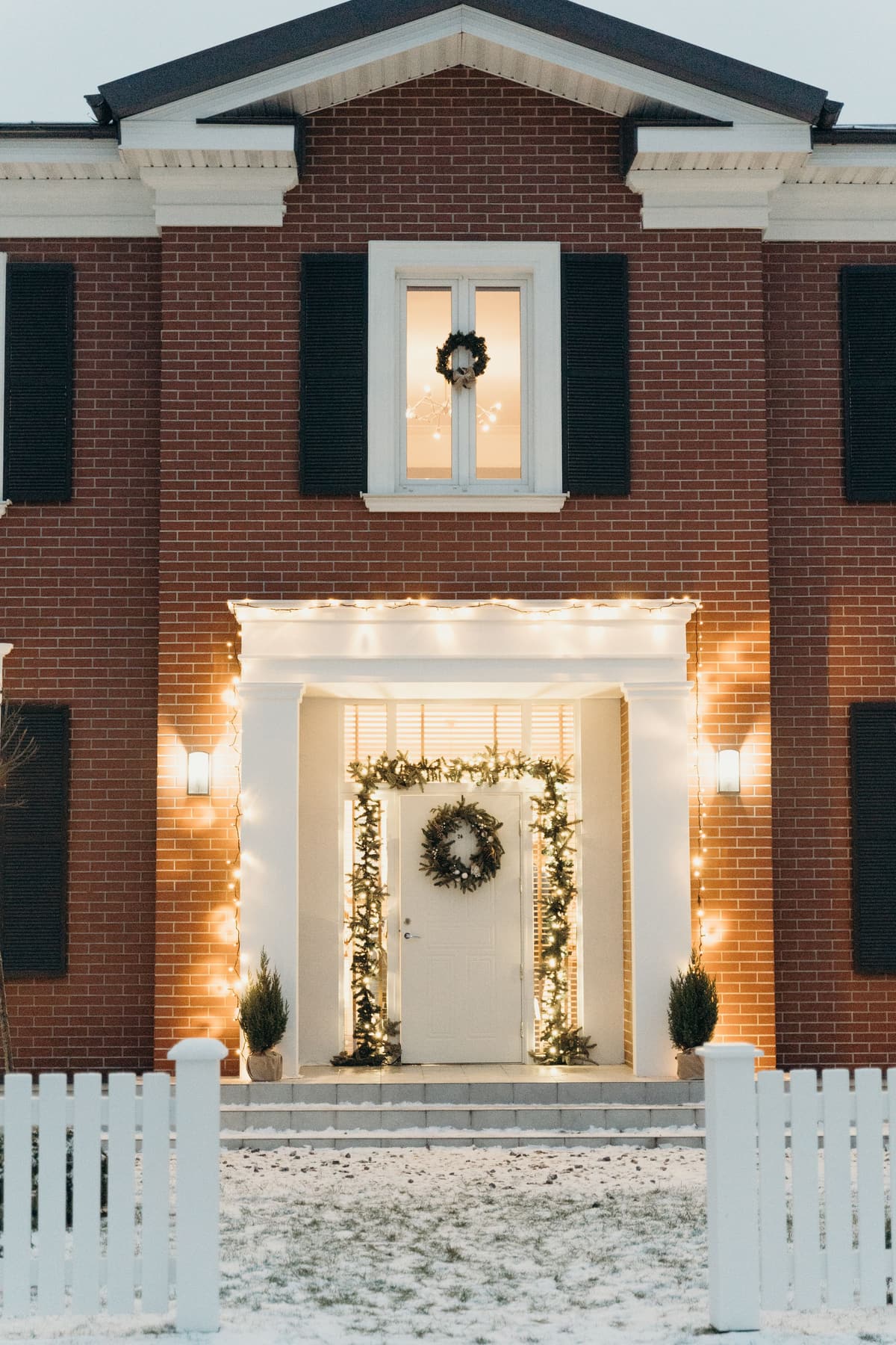 A symmetrical front entry at night with a wreath on the door, lit garland around the frame and warm white lights along the portico